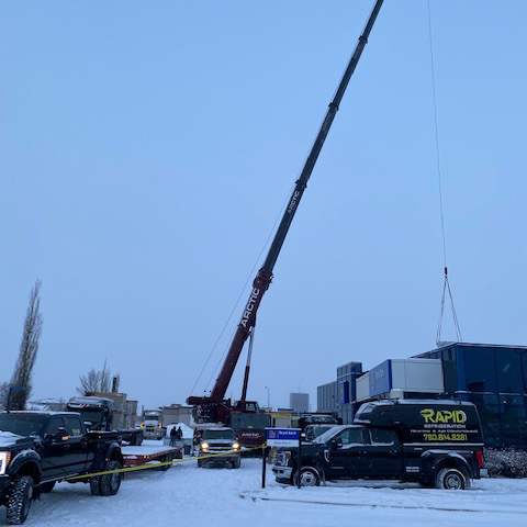 Crane lifting HVAC equipment on to an RBC Bank building in Grade Prairie. A Rapid Refrigeration truck and several other trucks are parked in front of the crane